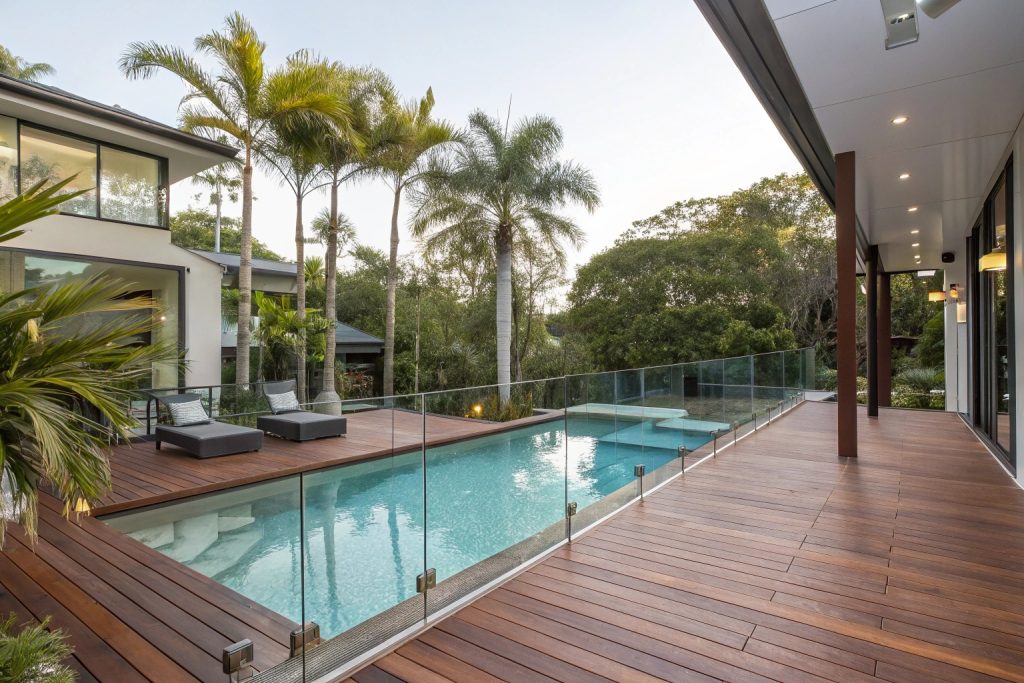 Timber deck wrapped around swimming pool in Broadbeach backyard with glass safety barriers and built-in seating areas